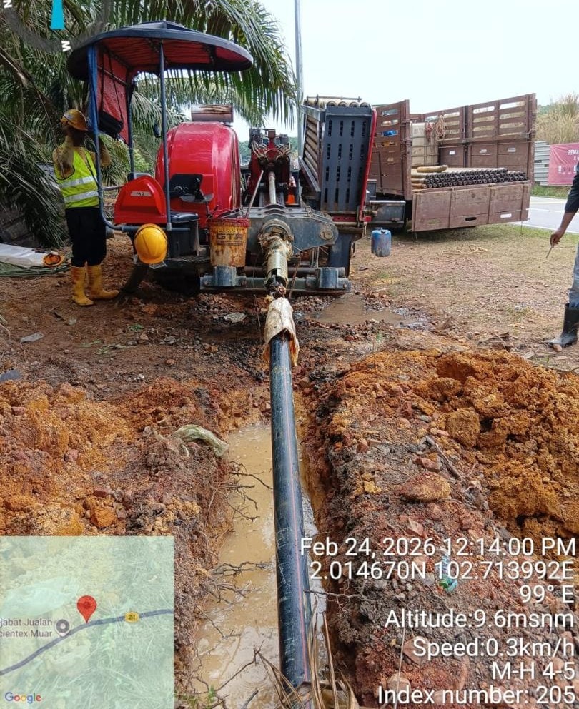 Aerial view of a deep straight trench cut through dry laterite soil alongside a highway, black corrugated duct visible at base of trench. No personnel visible. MH 20–19, Index 5470, Bukit Bakri, Johor (13 March 2026).