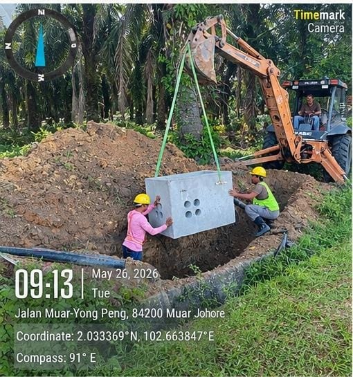 Two workers in hi-vis and hard hats guiding a precast concrete chamber being lifted by CASE excavator with green slings. Palm oil plantation backdrop. Jalan Muar–Yong Peng, 84200 Muar, Johor (26 May 2026).