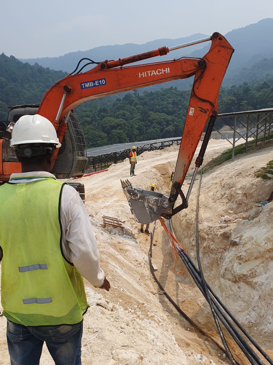 Hitachi excavator (TMB-E10) working alongside personnel at the cable trench, with solar panel structures and forested hillside in the background.